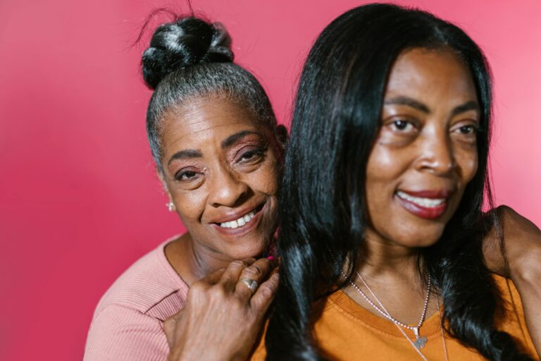 Close-up of two smiling African American women, a senior and an adult, against a pink backdrop.